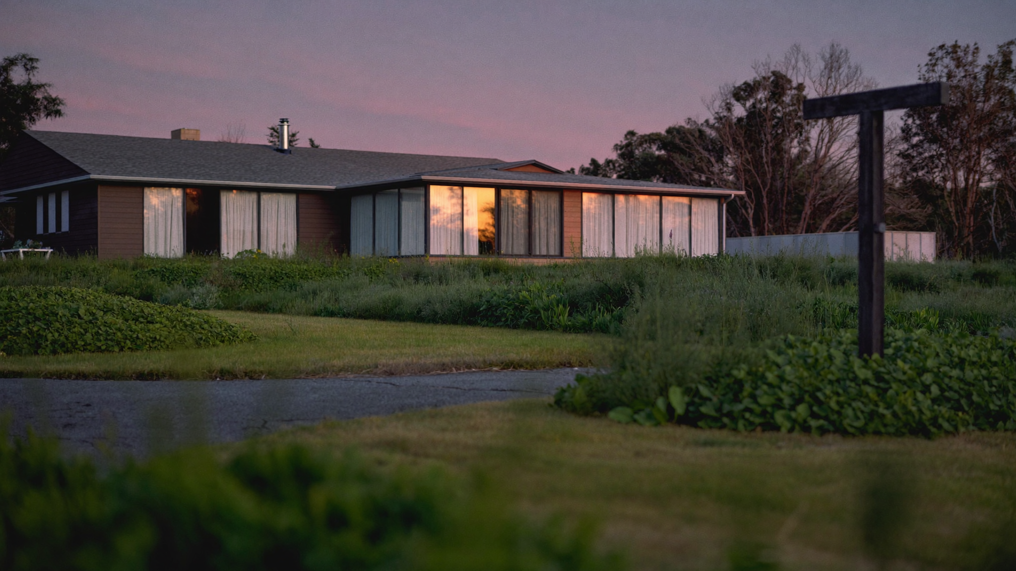 An empty vacation home at dusk with a for-sale sign in an overgrown yard