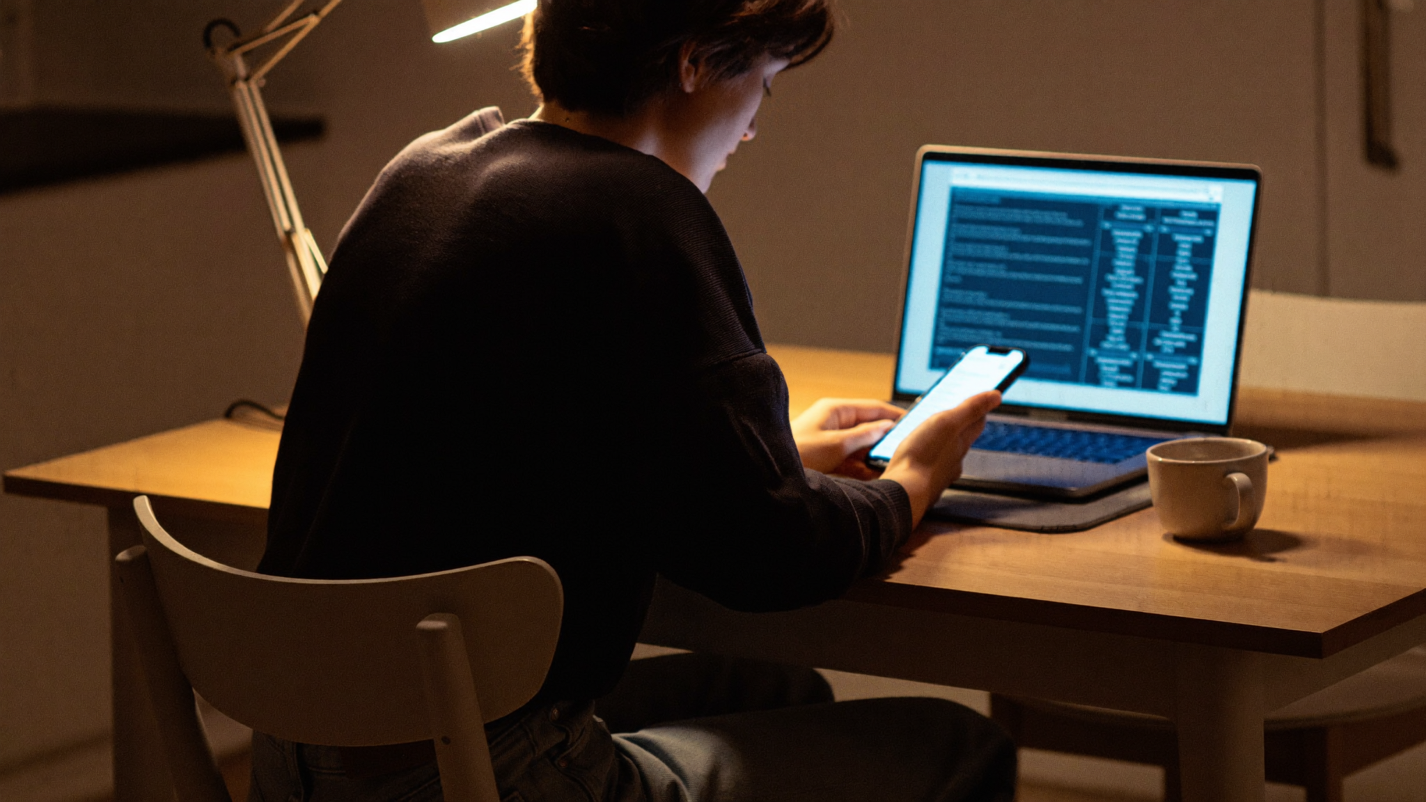 A person at a kitchen table at night studying a suspicious text message on a smartphone beside an open laptop