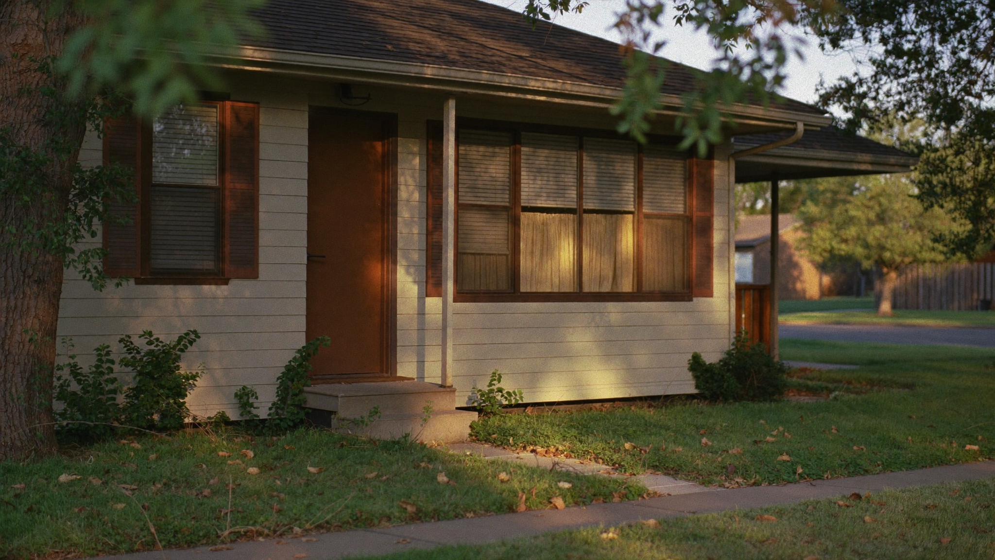 An older single-story family home on a quiet residential street, slightly overgrown yard, late afternoon light